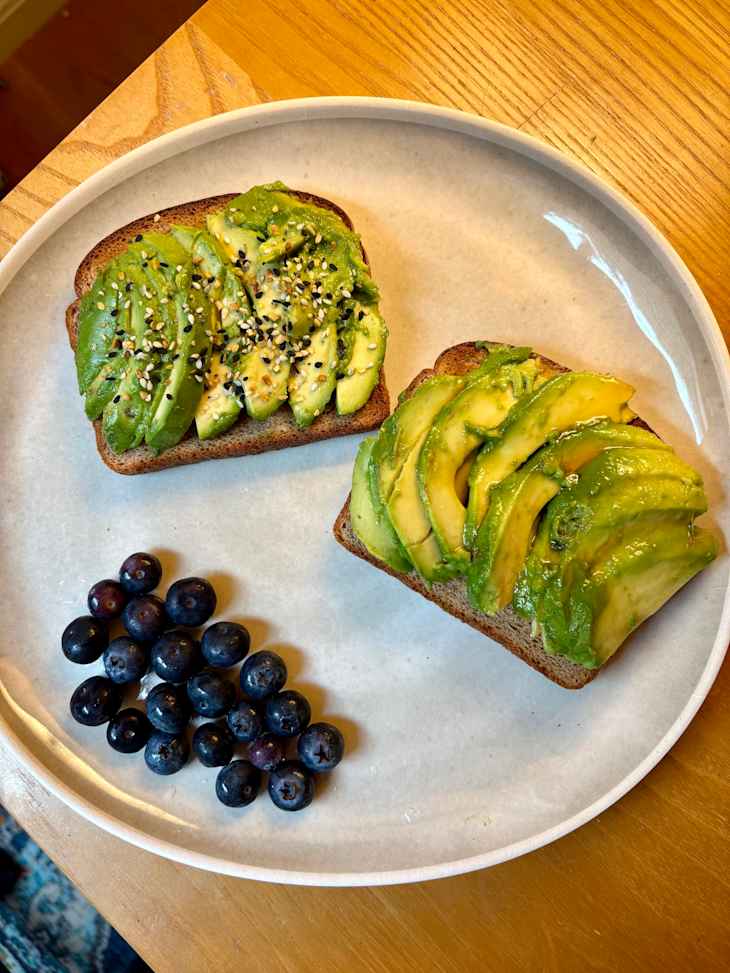 Two slices of avocado toast on whole grain bread, topped with sesame seeds, alongside a cluster of fresh blueberries.