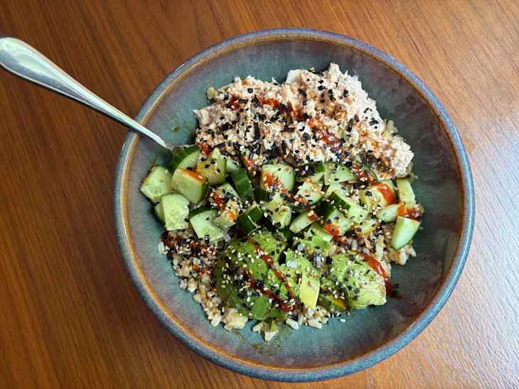 Bowl of rice topped with diced cucumbers, avocado, and tuna, garnished with sesame seeds and sauce.
