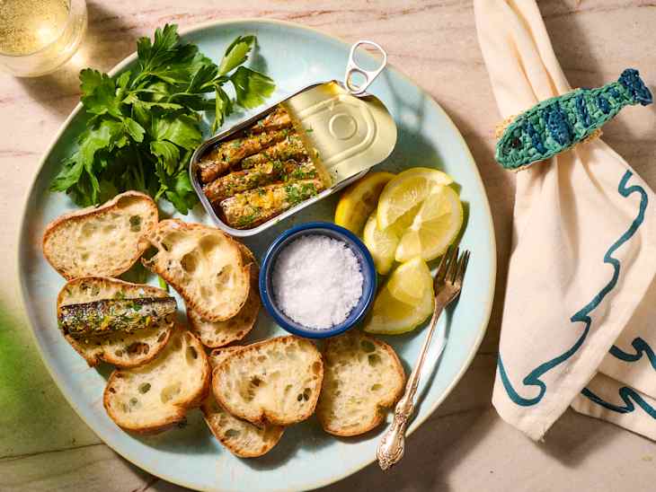 Sardines in a tin on a plate with toasted bread, lemon slices, fresh parsley, and a small bowl of salt.