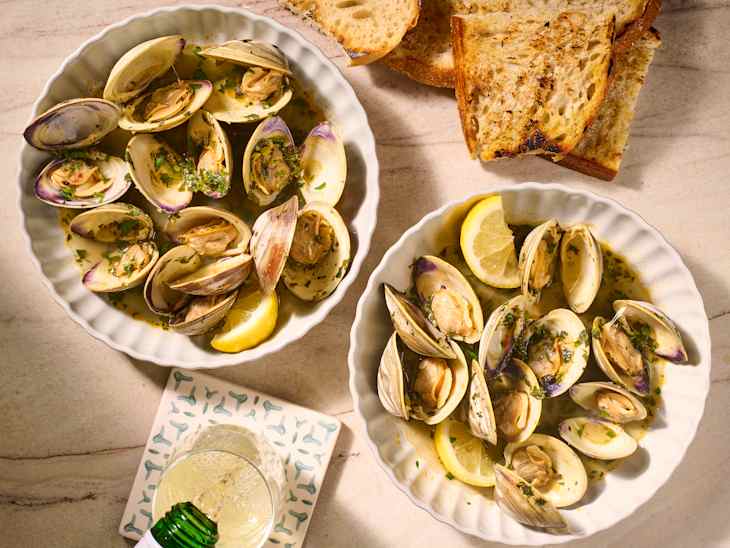 Two bowls of steamed clams garnished with herbs and lemon slices, accompanied by toasted bread and a glass of drink.