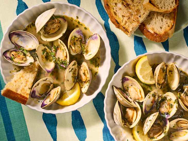 Clams in broth with herbs and lemon slices, served with toasted bread on a striped tablecloth.