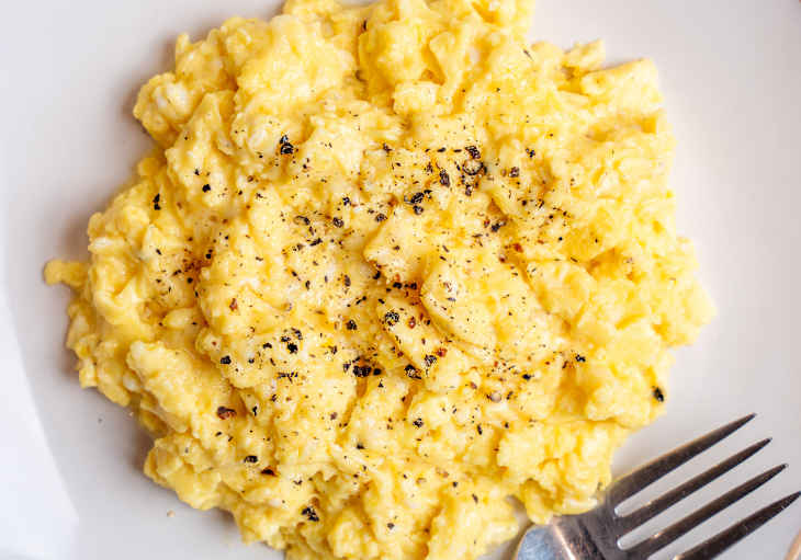 Fluffy scrambled eggs seasoned with black pepper, served in a white bowl with a fork.