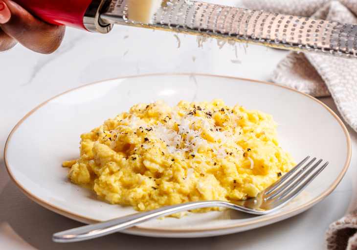 Creamy scrambled eggs topped with grated cheese and black pepper on a white plate, with a fork beside it.