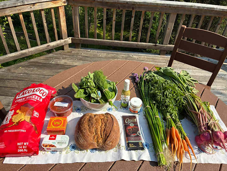 A variety of fresh vegetables, herbs, and snacks on a table, including carrots, greens, and a loaf of bread.
