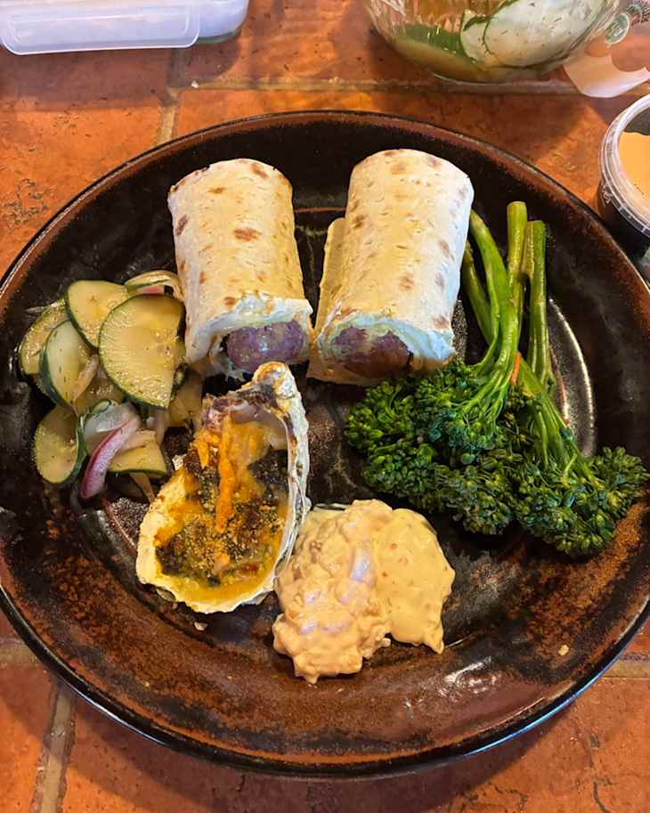 Two wrapped sausages on a plate with sautéed zucchini, broccolini, a baked oyster, and a creamy dipping sauce.