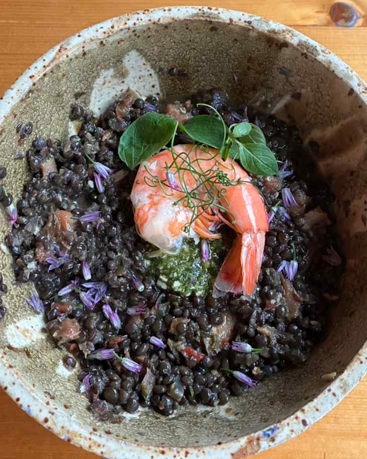 A bowl of black lentils topped with a large shrimp, green herbs, and purple edible flowers.
