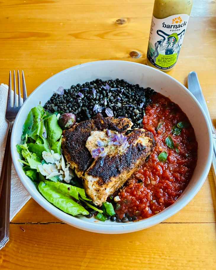 Bowl of black lentils topped with grilled fish, tomato sauce, greens, and edible flowers, with a bottle of sauce nearby.
