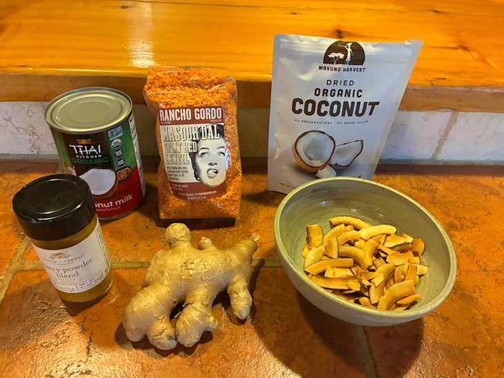 Canned coconut milk, red lentils, dried coconut, ginger root, and curry powder on a wooden countertop.