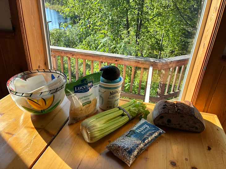 A bowl with napkins, parmesan cheese, celery, an avocado, navy beans, and a loaf of bread on a wooden table by a window.