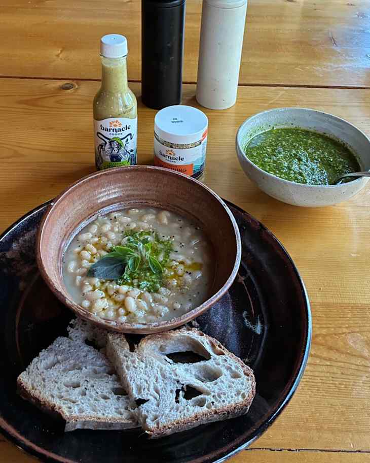 Bowl of creamy soup with white beans, garnished with herbs, served with slices of bread and condiments on a wooden table.