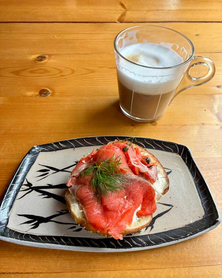 A plate with a toasted bagel topped with smoked salmon, dill, and capers, alongside a glass of latte.