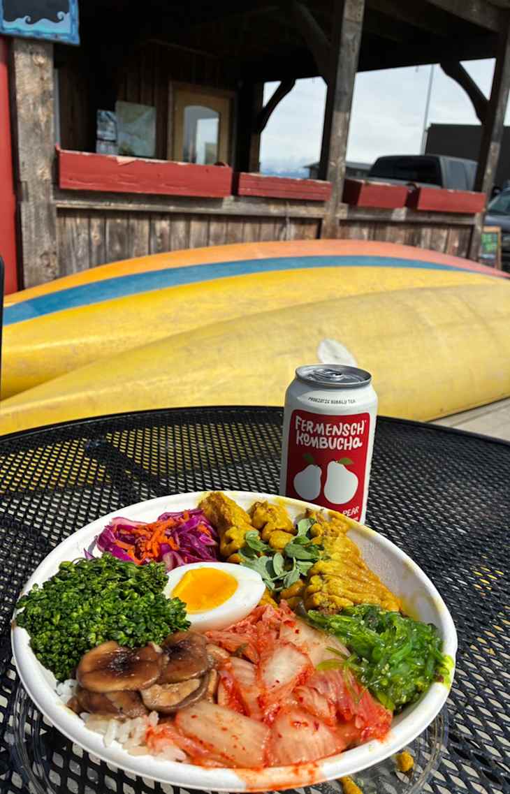 Bowl of rice topped with kimchi, mushrooms, greens, a soft-boiled egg, and a can of kombucha on a table.