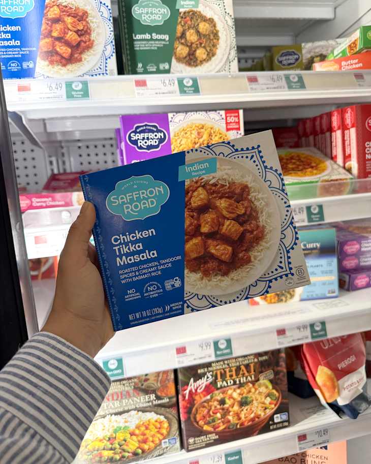 Frozen food aisle featuring a hand holding a Chicken Tikka Masala box with rice, surrounded by various Indian meal options.