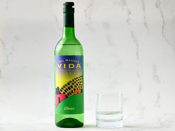 Green bottle of Del Maguey Vida Mezcal next to a clear glass with a small amount of liquid, set against a light marble background.