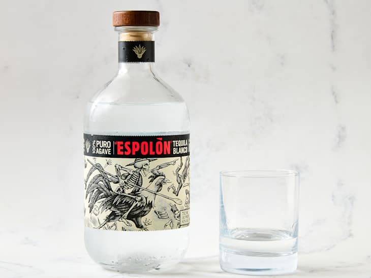 Clear bottle of Espolón Tequila Blanco with a wooden cap, next to an empty glass on a marble surface.
