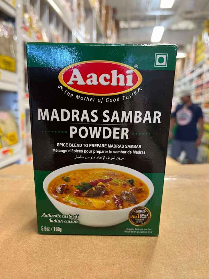 Box of Aachi Madras Sambar Powder with a bowl of sambar featuring spices, displayed in a grocery aisle.