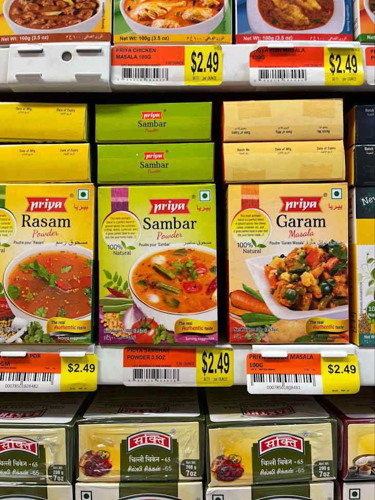 Various Priya spice packets on a store shelf, including Rasam, Sambhar, and Garam Masala, all priced at $2.49.
