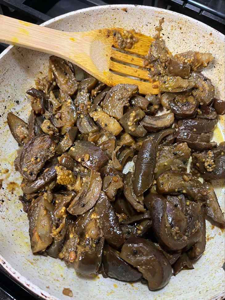 Sautéed eggplant pieces in a pan with a wooden spatula, garnished with sesame seeds and spices.
