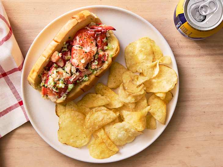 Lobster roll filled with lobster meat and vegetables, served with potato chips and a can of soda.