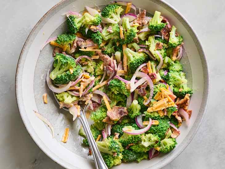 Bowl of broccoli salad with red onion, bacon bits, and shredded cheese, garnished with crispy toppings.