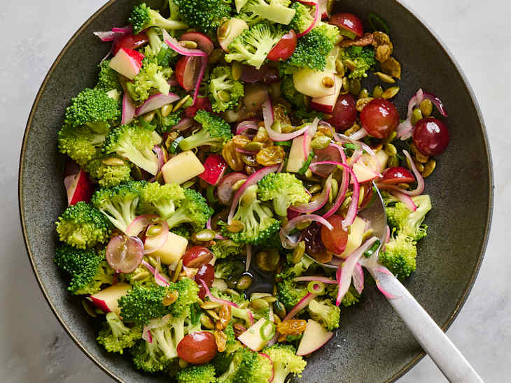 A vibrant salad featuring broccoli, red apples, grapes, and pumpkin seeds, garnished with red onion slices.