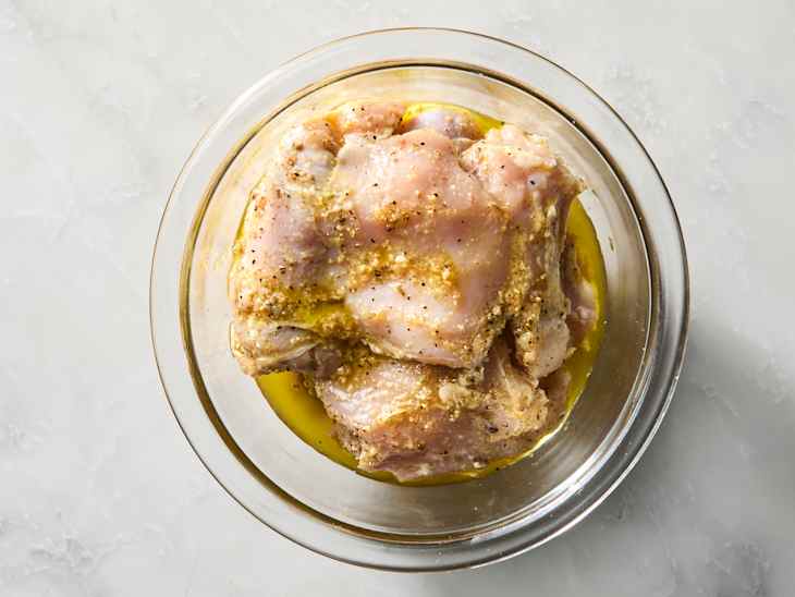 Marinated chicken thighs in a glass bowl with olive oil and spices on a light surface.