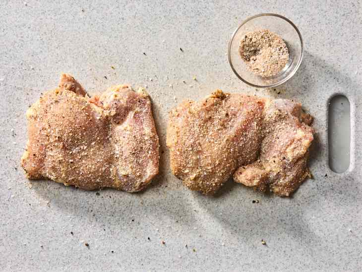 Two seasoned chicken thighs on a cutting board with a small bowl of spice mix nearby.