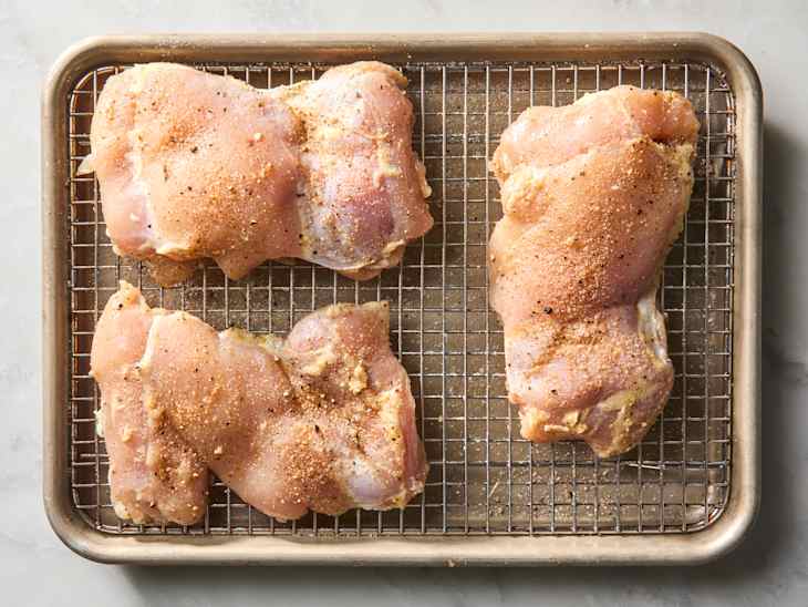 Raw chicken thighs seasoned with spices on a wire rack in a baking tray.