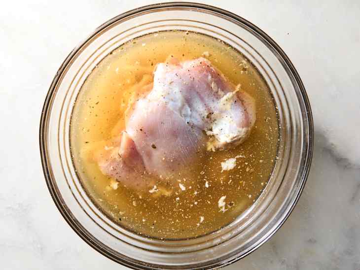 Raw chicken breast submerged in seasoned broth in a clear glass bowl on a marble surface.