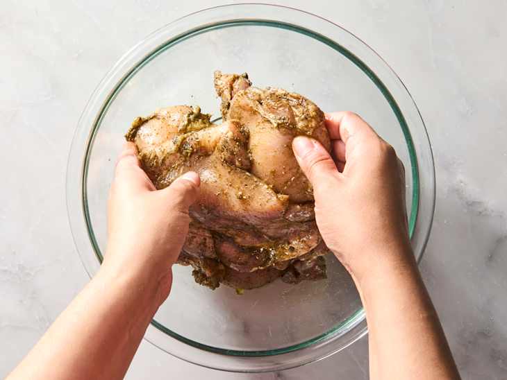 Marinated chicken being handled in a glass bowl, with hands applying seasoning.
