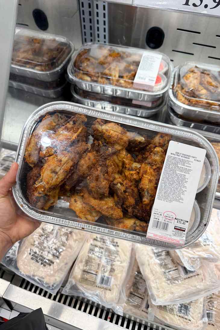 A hand holding a tray of garlic seasoned chicken wings with dipping sauces, displayed in a refrigerated section.