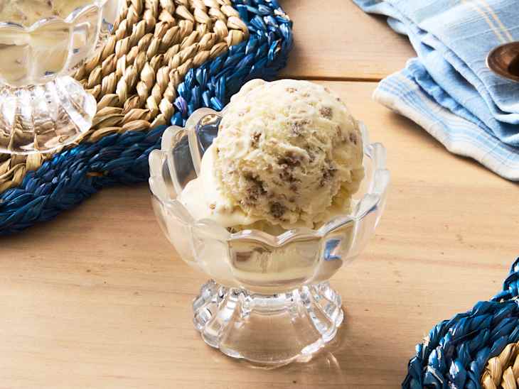 Scoop of ice cream with chocolate chunks in a clear glass dish, placed on a wooden table with woven placemats.
