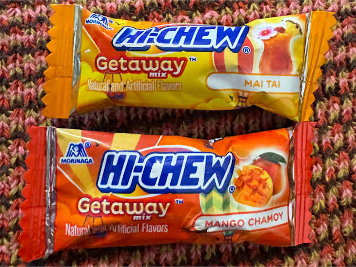 Two Hi-Chew candy wrappers, one labeled "Mai Tai" and the other "Mango Chamoy," on a textured pink and brown background.