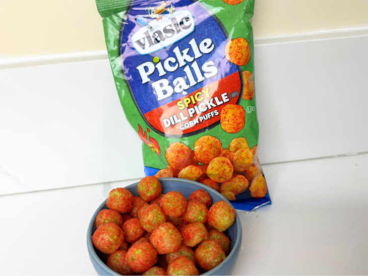 Spicy dill pickle corn puffs in a blue bowl next to an open bag on a white surface.