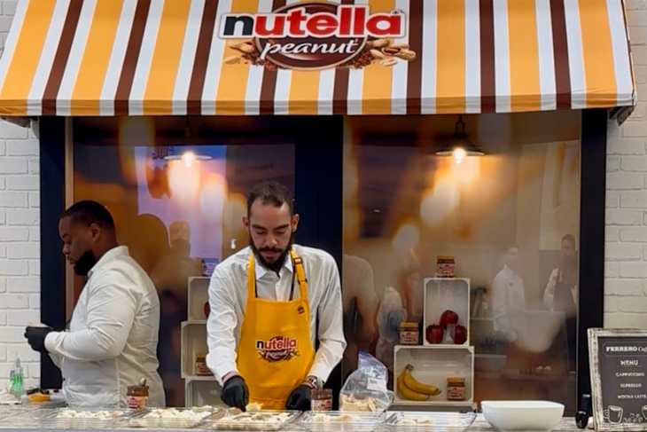 A man in a yellow apron prepares food at a Nutella-themed booth, with jars and fruits displayed behind him.