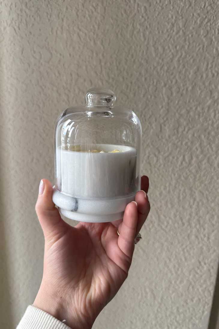 A hand holding a glass cloche over a white candle with a marble base, against a textured wall.