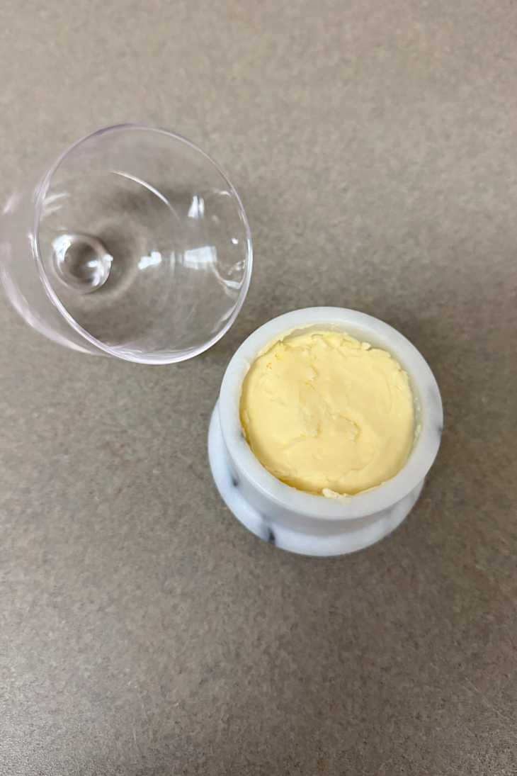 A marble container with yellow butter or cream, next to a clear glass lid on a beige surface.