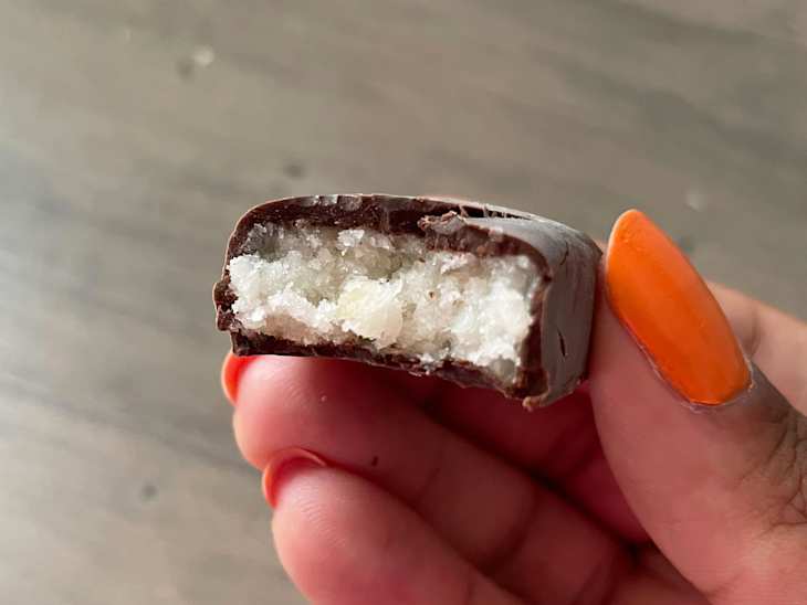 A hand holding a chocolate-covered candy with a bite taken out, revealing a white, textured filling.