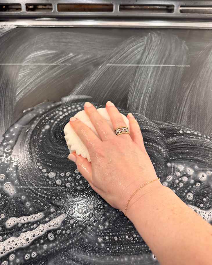 A hand with a ring scrubbing a soapy black surface with a sponge, creating bubbles and streaks.