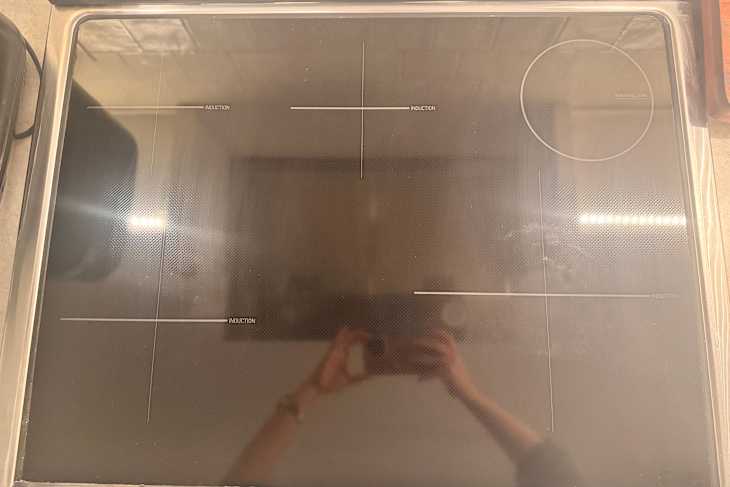 Induction cooktop with four cooking zones, including a warming zone, reflecting a hand holding a camera.