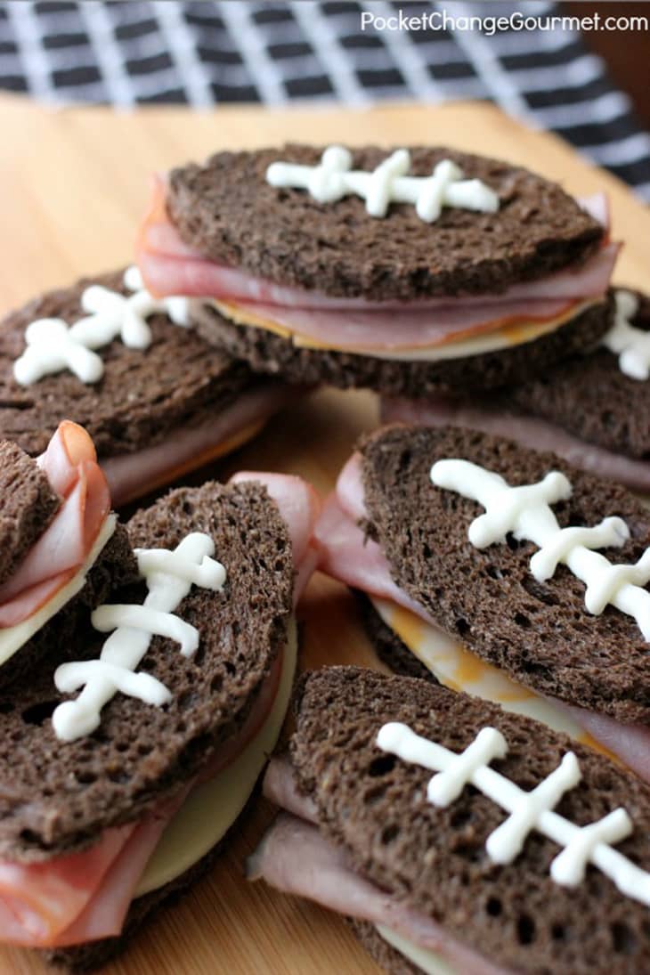 Football-shaped sandwiches with dark bread, ham, cheese, and cream cheese laces on a wooden board.