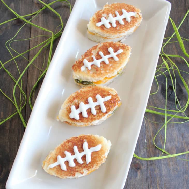 Four football-shaped grilled cheese sandwiches with white icing laces on a rectangular white plate.
