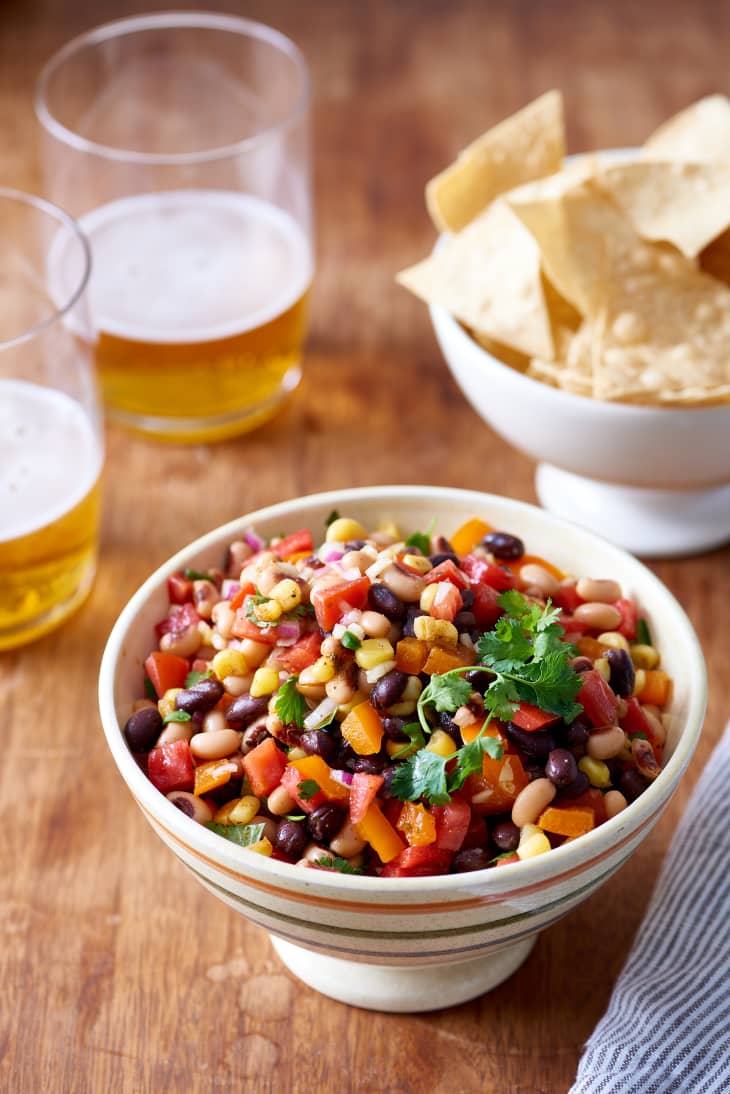 Bowl of colorful bean salad with corn, tomatoes, and cilantro, next to glasses of beer and a bowl of tortilla chips.