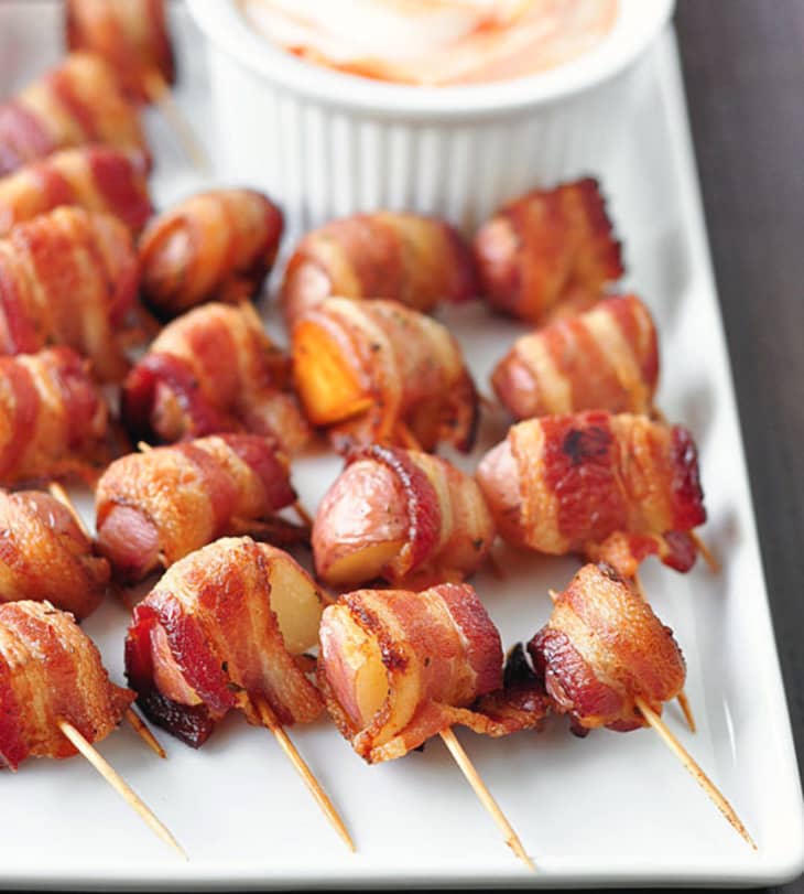 Bacon-wrapped potatoes on skewers served on a white platter with a dipping sauce in a ramekin.