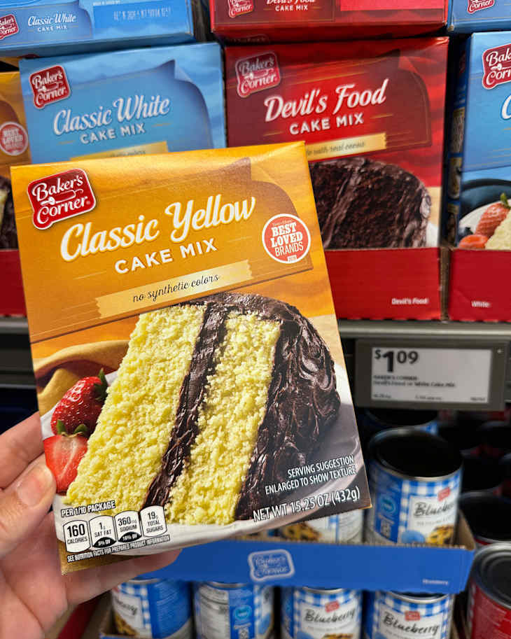Box of Baker's Corner Classic Yellow Cake Mix with a slice showing yellow cake and chocolate frosting, surrounded by other cake mixes.