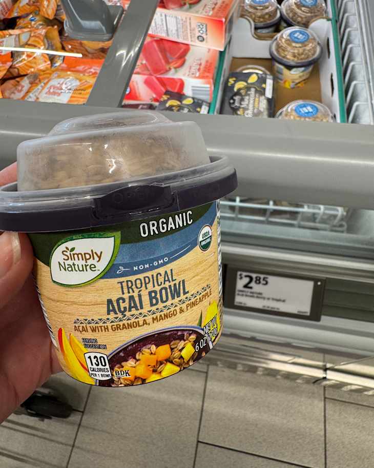 A hand holding a Tropical Acai Bowl with granola, mango, and pineapple in a grocery store freezer aisle.