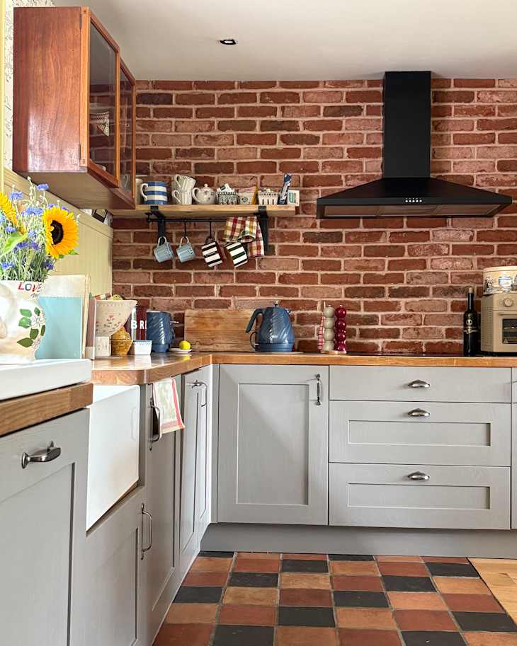 Modern kitchen with gray cabinets, brick wall, wooden countertop, blue kettle, and colorful dishware on shelves.