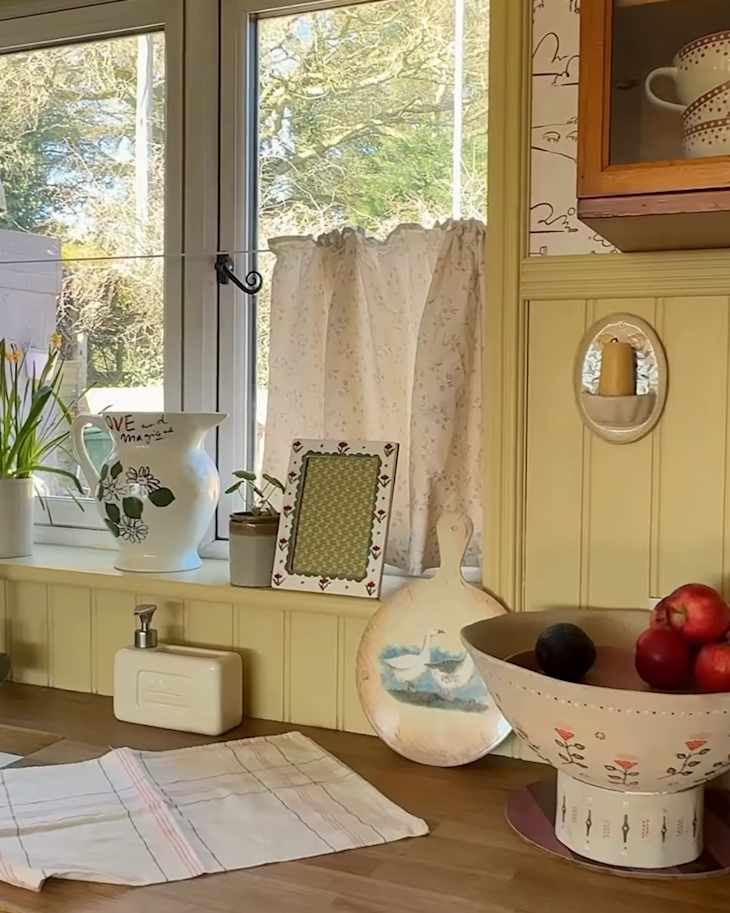 Cozy kitchen corner with a wooden countertop, decorative bowl of apples, potted plants, and a floral curtain.