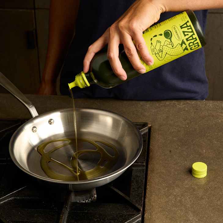 A person pouring Graza oil from a bottle into a stainless steel frying pan on a stovetop.