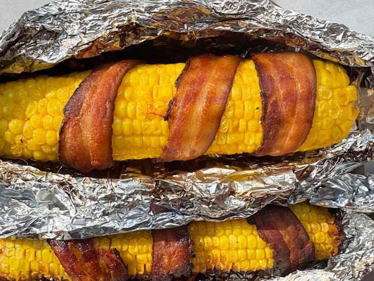 Grilled corn on the cob wrapped in bacon, nestled in aluminum foil.